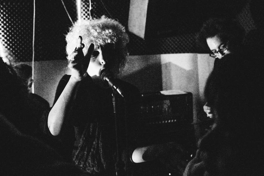 A vocalista da banda Inês é Morta, Camila, aparece cantando no microfone na frente do grupo em uma foto em preto e branco. Ela tem o cabelo loiro bem claro e cacheado em formato redondo, estilo anos 70